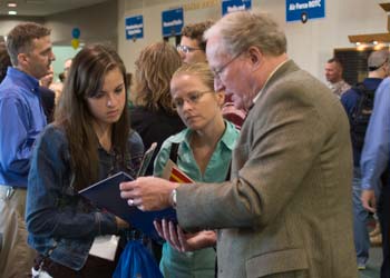 A young woman assists a girl with her homework at a small table in a room full of students.