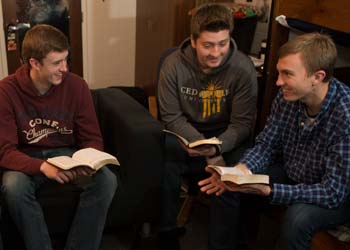 Three male students gather in a softly lit dorm room to study the Bible