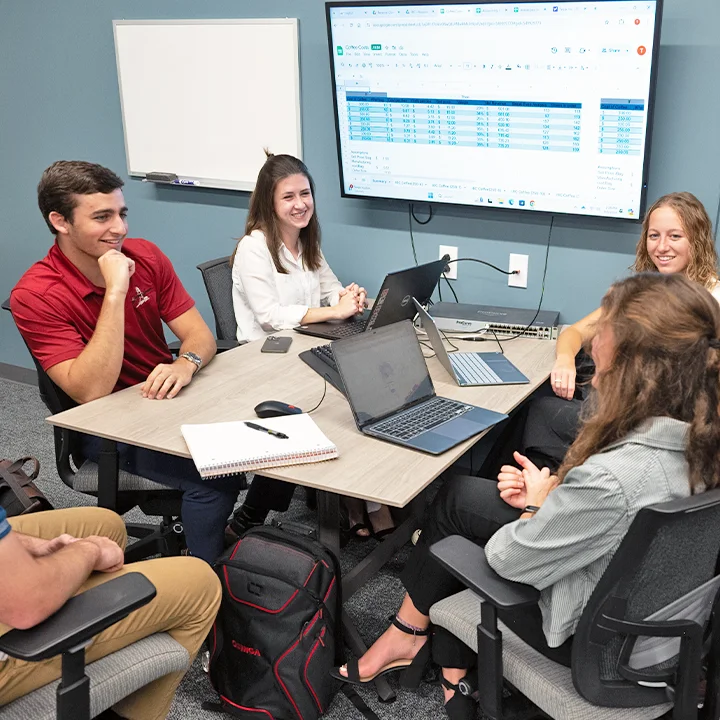 Small group of students gathered at table with laptops and notebooks.