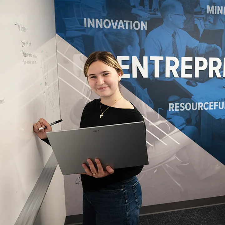 Student smiling while holding laptop in one hand and writing on whiteboard with the other hand.