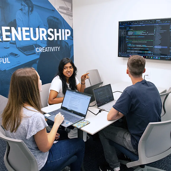 Three students gathered at table with laptops looking at computer code on a TV screen.
