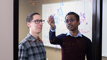 Two male students performing math equations on a glass wall.