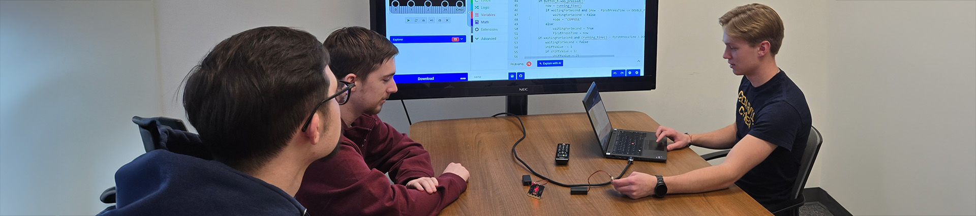 Three young men seated at table doing work on a large computer monitor.