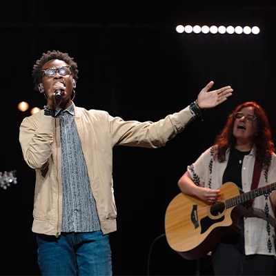 Student singing into microphone onstage and lifting hand in worship.