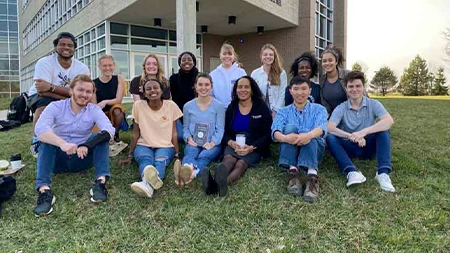 Group of 13 students and faculty member smiling outside, with some holding a book.