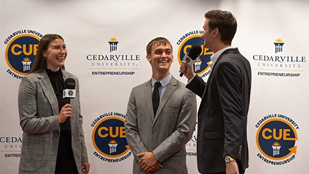 Two students interviewing another student by a backdrop that says 