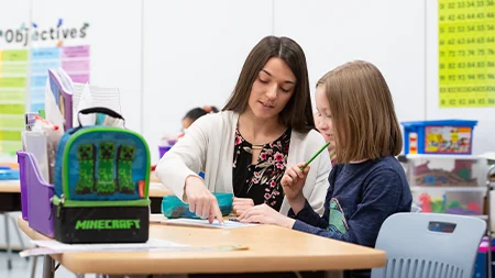 Student teacher helping elementary school student at her desk.