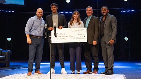 Two students holding a check and smiling with panel of three judges onstage.