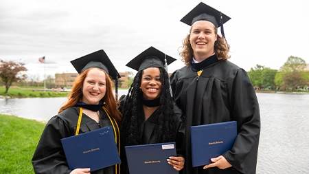Three college students in graduation regalia smiling by lake
