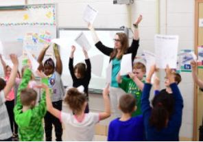 education major in classroom with young students with hands raised and papers in hand