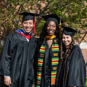 Three students in college graduation caps and gowns posing for picture outside