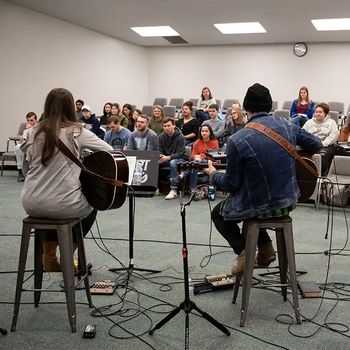 Two people playing guitars in room of people.